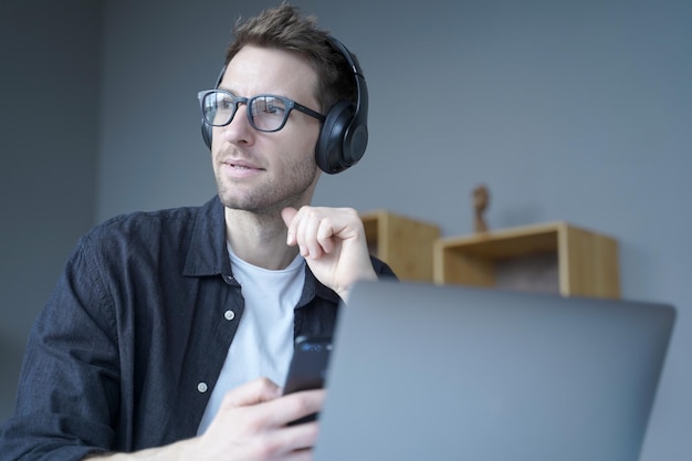 Thoughtful young businessman in headphones holds smartphone while sits at workplace at home