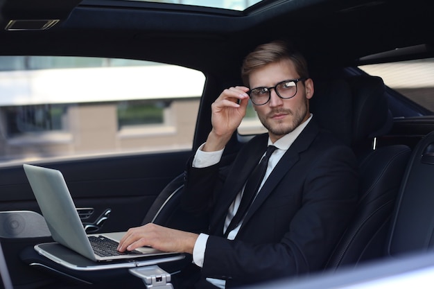 Thoughtful young businessman keeping hand on glasses while sitting in the lux car and using his laptop.