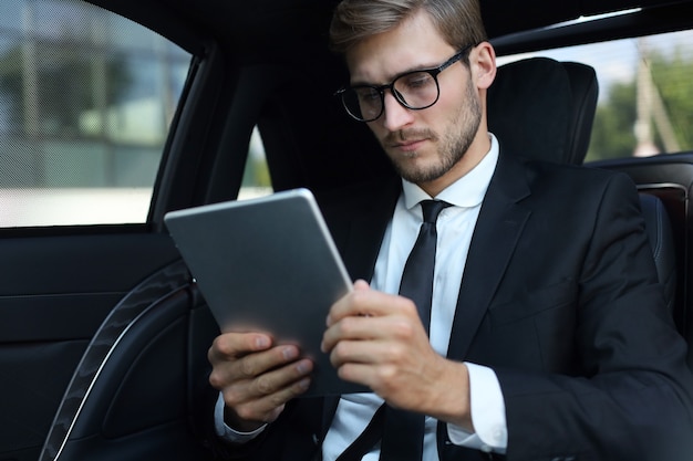 Thoughtful young businessman sitting in the luxe car and using his tablet.