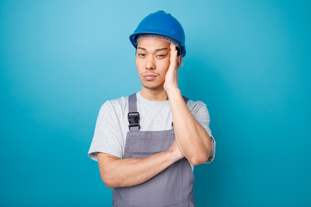 Thoughtful young construction worker wearing safety helmet and uniform keeping hand on head 