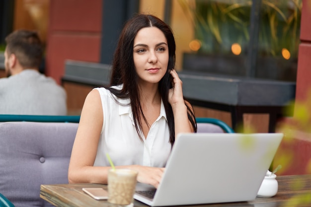 Thoughtful young female in elegant blouse, looks pensively into distnace, works on laptop computer, drinks aromatic coffee