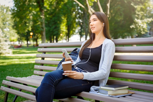 Thoughtful young female student in headphones. Relaxing and listening to music, while drinking coffee in the park. Lifestyle and technology concept