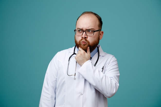 Thoughtful young male doctor wearing glasses medical coat and stethoscope around his neck keeping hand on chin looking at side isolated on blue background