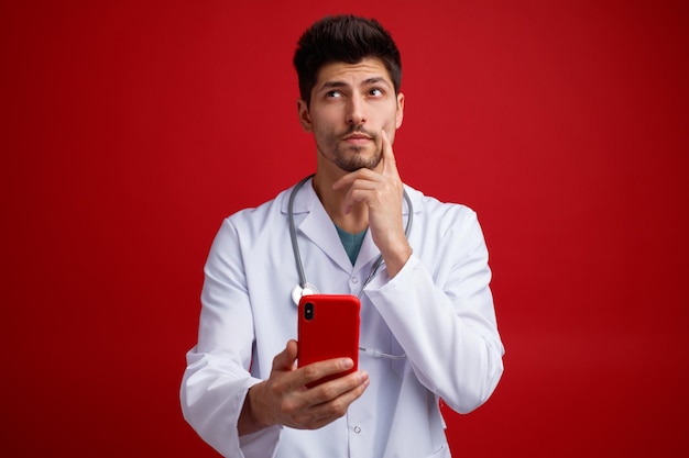 Thoughtful young male doctor wearing medical uniform and stethoscope around his neck holding mobile phone keeping hand on chin looking up isolated on red background