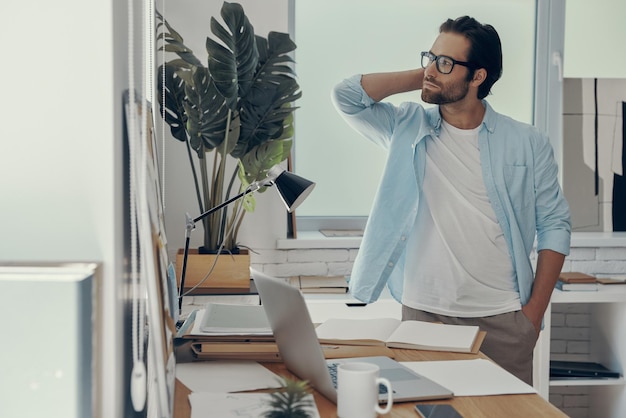 Thoughtful young man looking away while standing near his working place in office