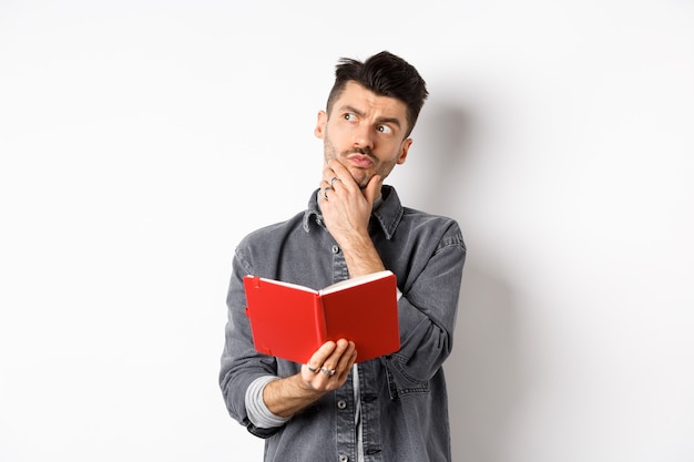 Thoughtful young man plan his schedule, looking aside pensive, holding opened planner or journal, standing on white background.