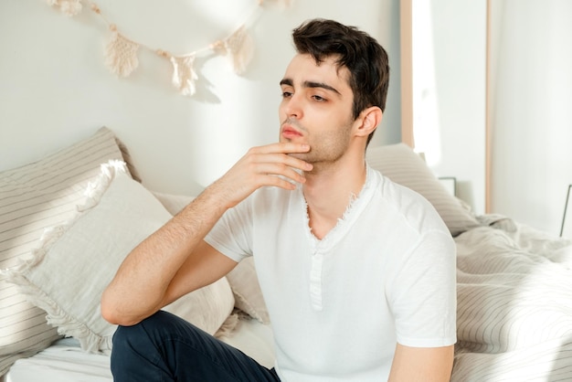 Thoughtful young man sitting on the bed in the bedroom touching his chin and thinking Thoughtful mature man resting on bed at home feeling sad thinking about future