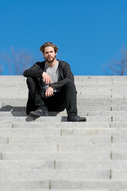 Thoughtful young man sitting on a flight of steps staring into the distance with a serious expression against a blue sky background