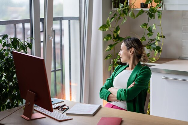 Thoughtful young tired business woman looking at window while sitting at workplace of home office