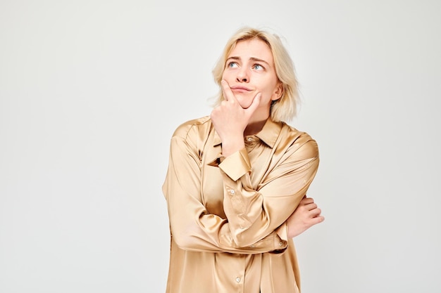 Thoughtful young woman in beige blouse looking upwards hand on chin isolated on white background