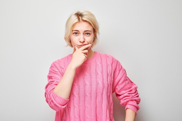 Thoughtful young woman in pink sweater with hand on chin looking at camera on light grey background