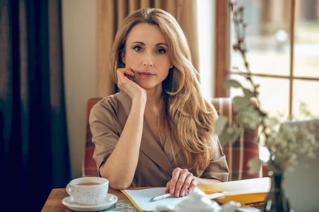 In thoughts. Blonde long-haired woman sitting at the table and looking thoughtful