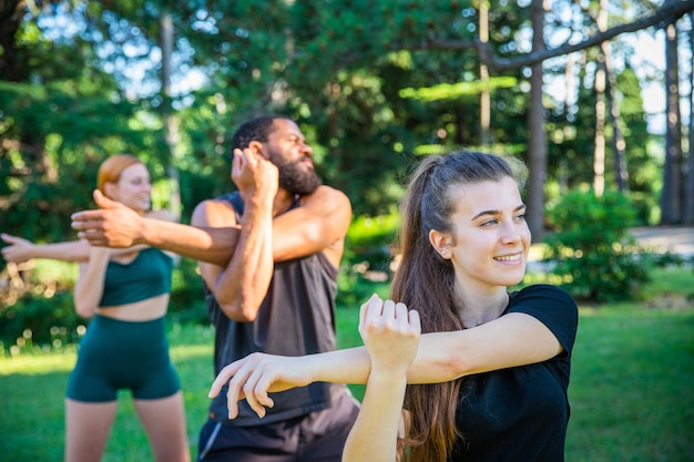 Three athletes train and perform an arm stretch exercise outdoors in a public park