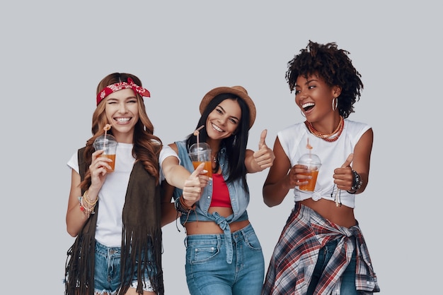 Three attractive young women keeping thumbs up and smiling while standing against grey background