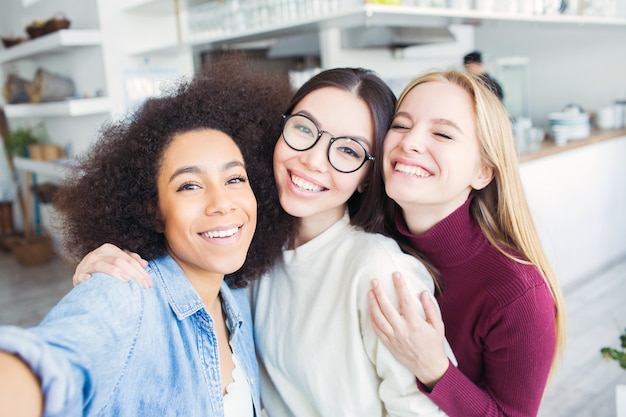 Three beautiful friends are taking selfie together. There are afro american girl, one brunette and one blonde. THey are looking and smiling. Girls are very cheerful.