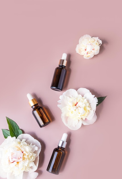 Three bottles with a dropper cosmetic product or serum on a pastel background with the heads of peonies natural cosmetics Top view Flat lay