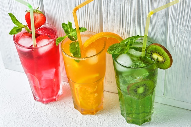 Three bright colored delicious cold lemonade carafes of kiwi and orange and strawberry with mint against the surface of a wooden white wall