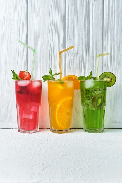 Three bright colored delicious cold lemonade carafes of kiwi and orange and strawberry with mint against the surface of a wooden white wall