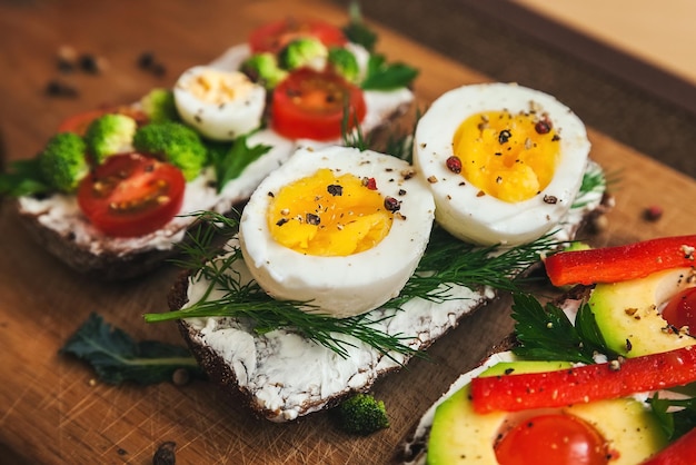 Three bruschettas on dark bread with curd cheese eggs vegetables spices and herbs on a wooden board