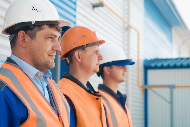 Three builders in helmets and vests stand in row Friendly construction team of Caucasian appearance Portraits of young workers at construction site on summer day