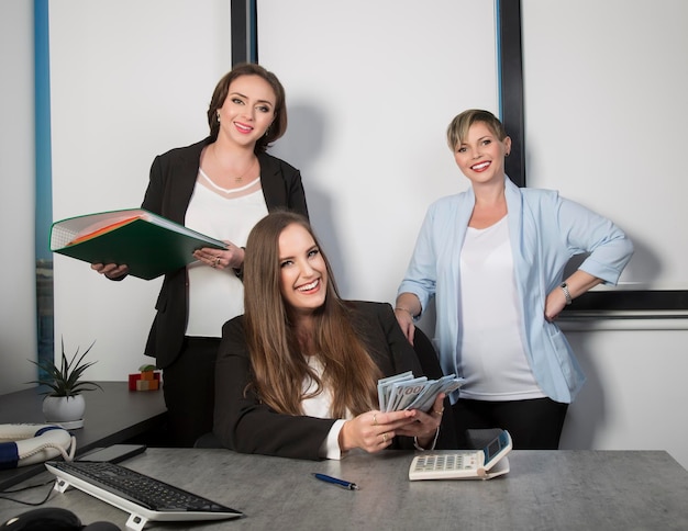 Three businesswoman at a work laughing while working at an office table Womans with cash money in dollar banknotes Female with long hair holding bundle dollars with calculator counting revenue