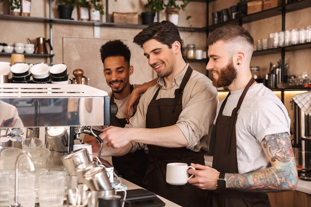 Three cheerful male baristas standing at the coffee shop counter indoors