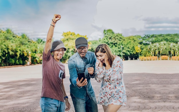 Three cheerful people looking at cell phone outdoors Excited teenage friends looking at cell phone screen on the street Three excited friends looking at the cell phone in the street