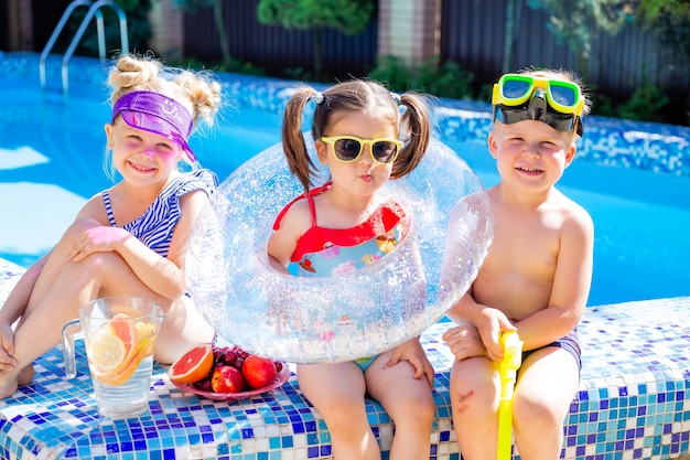 three children in the summer sit by the pool in sunglasses and drink lemonade