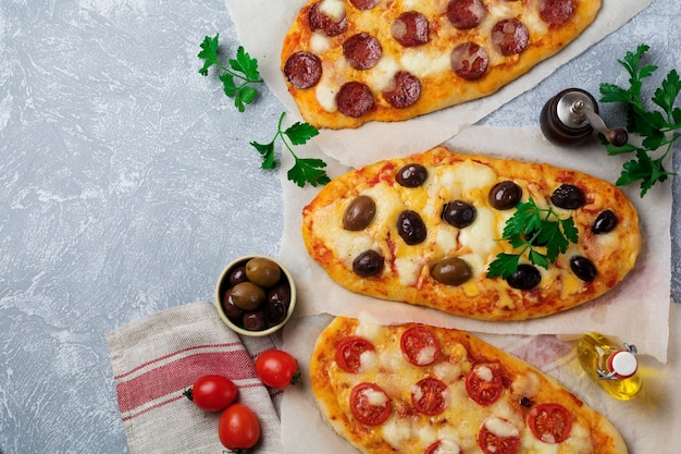 Three different oval pizza with olives, tomato and salami on a gray concrete.   