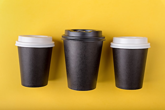 Three disposable paper black cups different sizes for takeaway coffee flat lay on yellow background