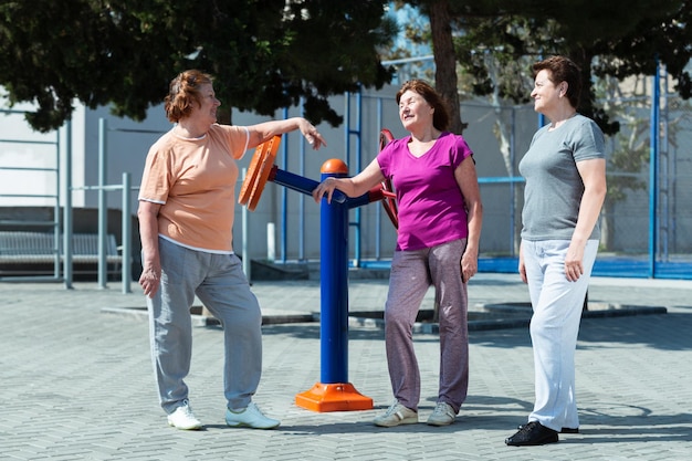 Three elderly woman on sport ground in a public park resting after training and talking