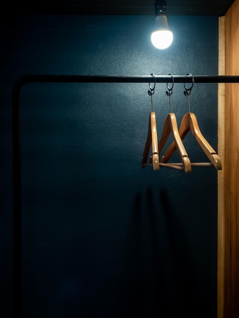 Three empty wooden clothes hangers without shirts or dress hanging on a black cloth rack with light bulb in the wood wardrobe on dark blue background in hotel room with copy space vertical style