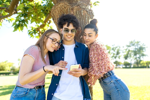 Three friends having fun using smartphone at park pointing and texting on display.