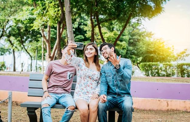 Three friends sitting on a bench taking a selfie Three teenage friends sitting on a bench taking selfies Gathering of three happy friends taking a selfie sitting on a bench