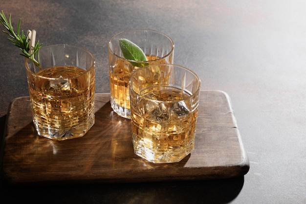 Three glasses of cold whiskey served on rocks with rosemary, lemon peels on brown. Copy space.