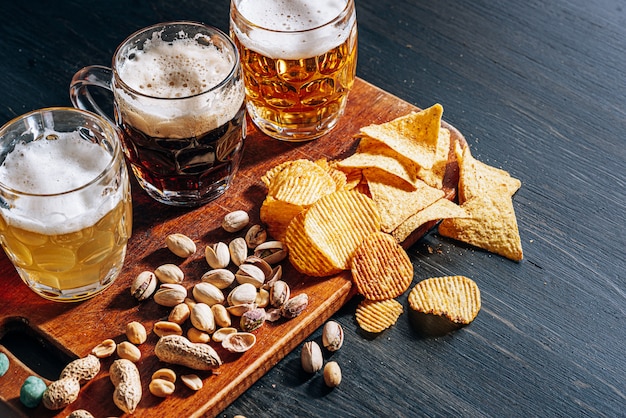 Three glasses of expensive craft beer, classic and unfiltered and dark in a glass on the table with a snack of peanut and pistachio chips and nachos