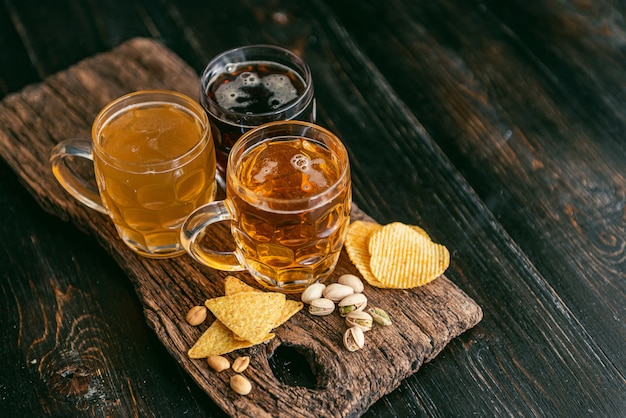 Three glasses of expensive craft beer, classic and unfiltered and dark in a glass on the table with a snack of peanut and pistachio chips and nachos