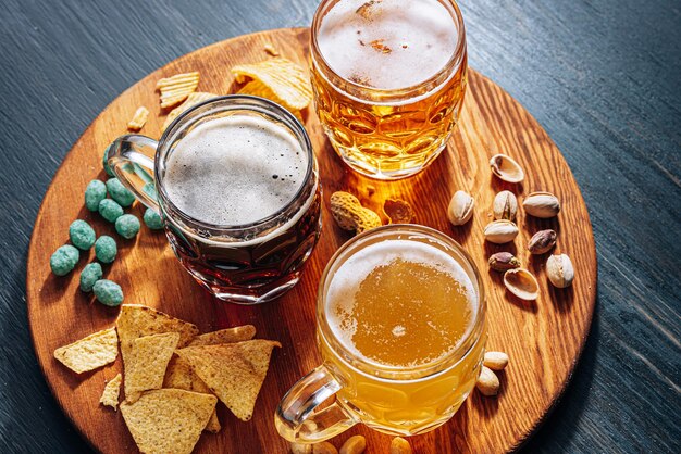 Three glasses of expensive craft beer, classic and unfiltered and dark in a glass on the table with a snack of peanut and pistachio chips and nachos