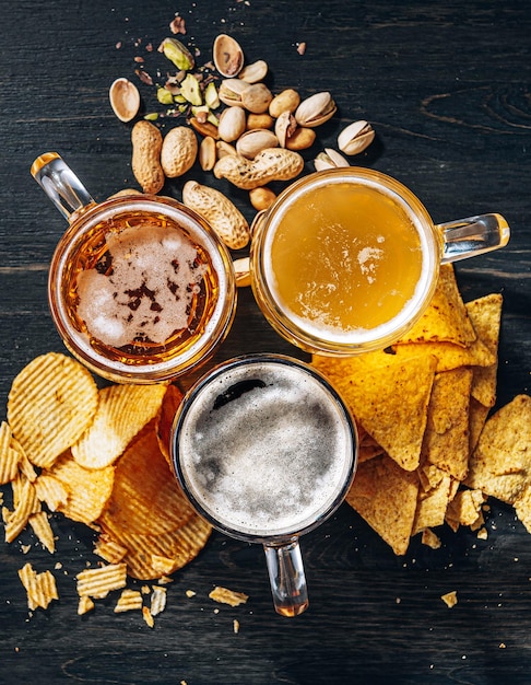 Three glasses of expensive craft beer, classic and unfiltered and dark in a glass on the table with a snack of peanut and pistachio chips and nachos