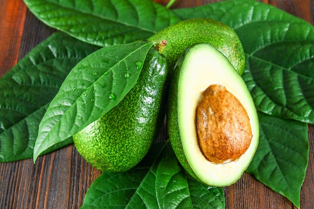 Three green raw ripe avocado fruits and a cut half with a bone with leaves on a brown wooden table.