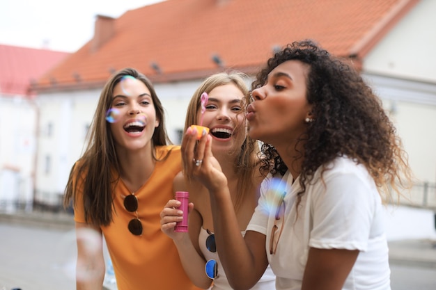 Three happy beautiful women blowing soap bubbles in city street.