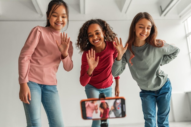 Three Happy Diverse Girls Waving Hands To Smartphone Indoors