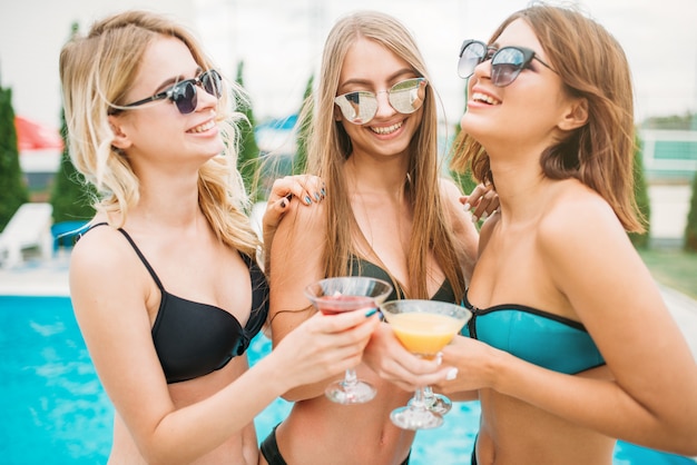 Three happy girls in swimsuits and sunglasses drinks cocktails and have fun near the swimming pool. Resort holidays. Tanned women on summer holidays