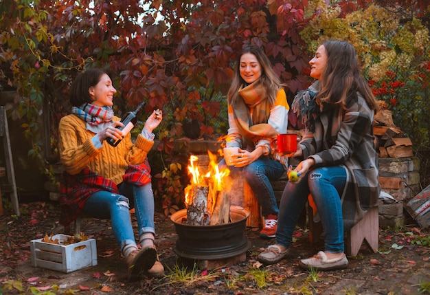 Three happy joyful best friends sitting outside in autumn garden by campfire with bottle of red wine