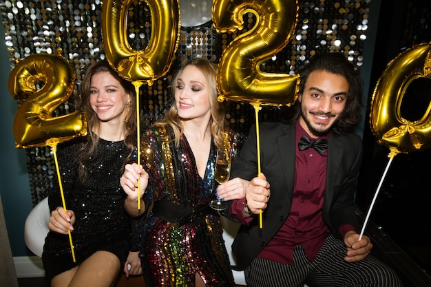 Three happy young friends holding golden inflatable numbers of the following year while having fun at party