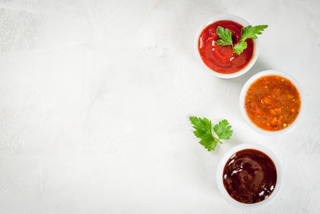 Three kinds of red tomato sauce on a white stone table: traditional classic ketchup, barbecue sauce, sweet and sour Chinese sauce. Top view copy space