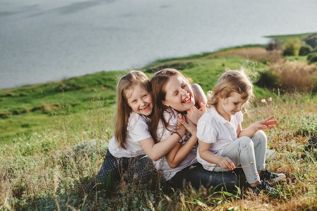 Three laughing girls in white sit in an embrace in the grass on the beach in summer