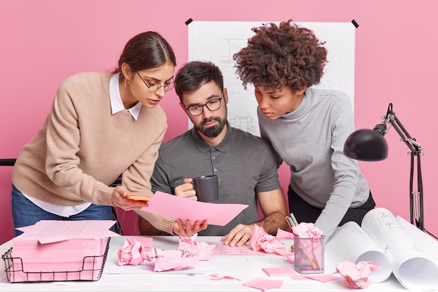 Three mixed race office coworkers collaborate on new building project look attentively in paper with information surrounded by sketches blueprints discuss ideas for illustation pose at coworking space