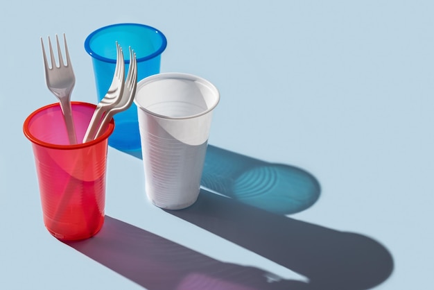 Three multi-colored plastic cups, cutlery on a blue background. Long shadows.