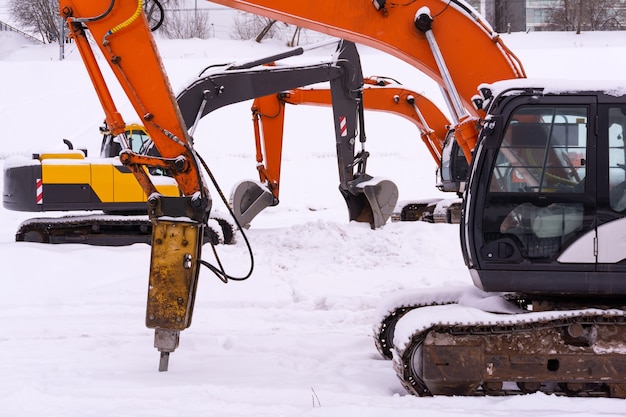 Three multicolored tracked excavators are parked on a snowy field, booms and sticks form an enfilade